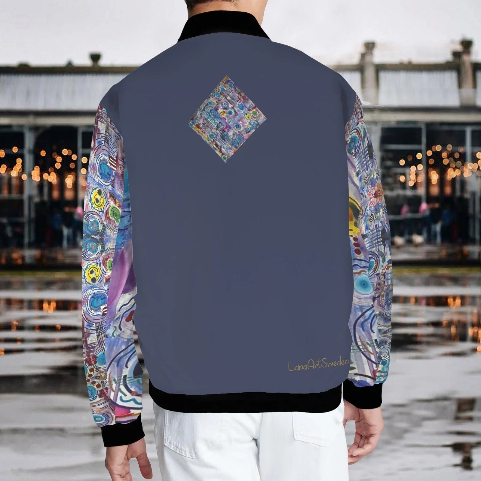 Back view of a deep blue bomber jacket with colorful circular patterns on sleeves and a diamond-shaped pattern on the upper back, worn by a person standing in an urban outdoor setting.