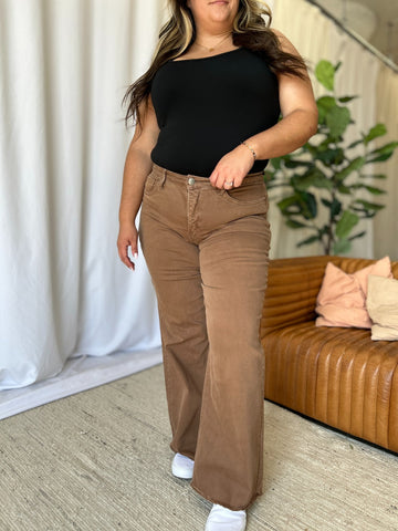 Woman wearing brown RFM Full Size High Rise Garment Dye Wide Leg Jeans with raw hem and black top indoors