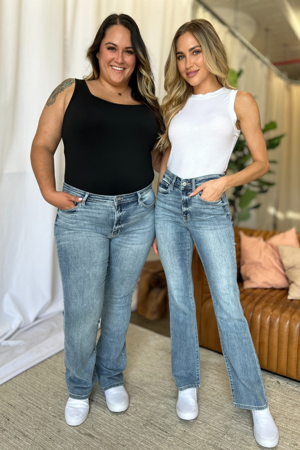 Two women wearing Judy Blue Full Size medium rise bootcut jeans in light washed denim with casual tops and white sneakers indoors