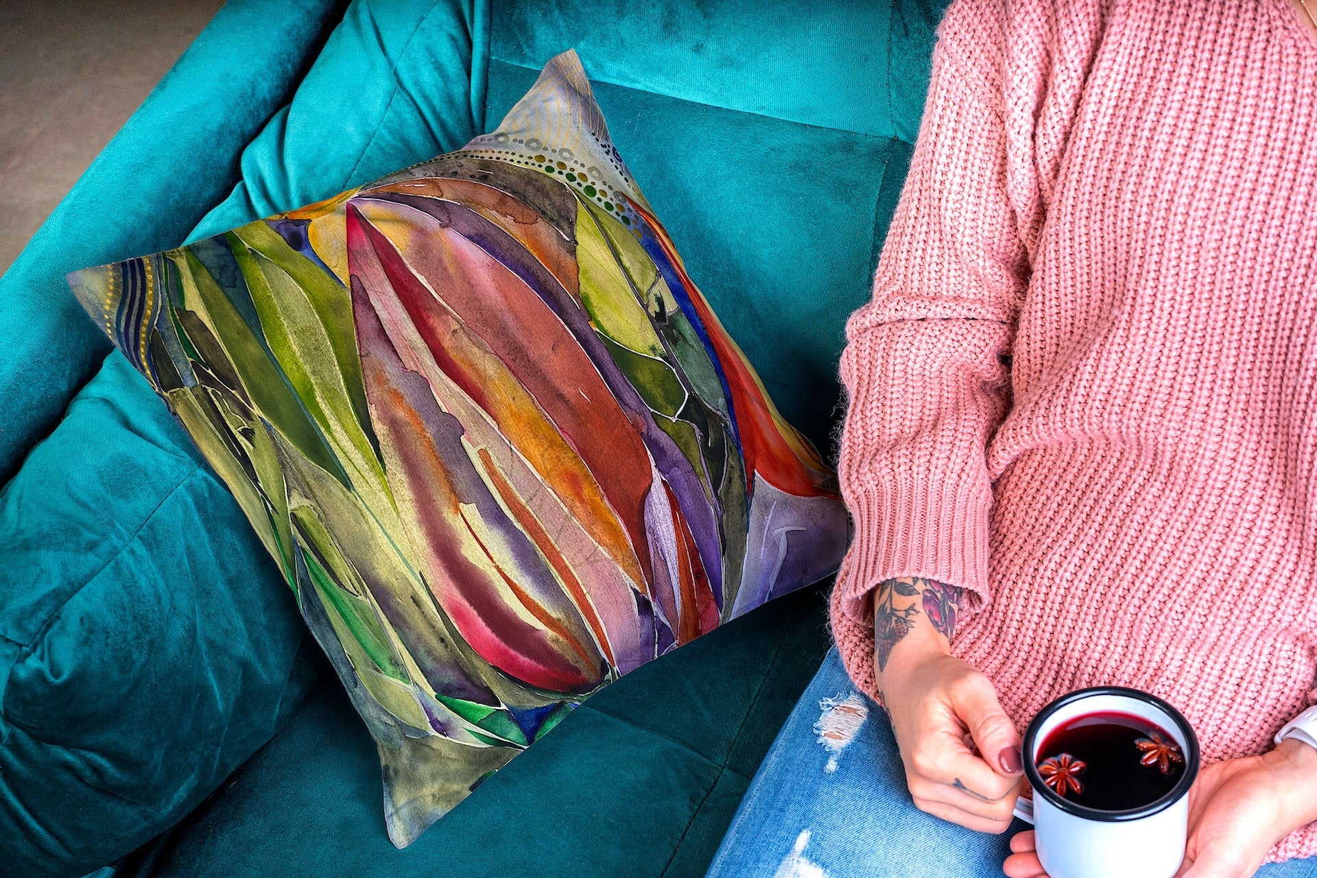 Banana Flower Pillow with vibrant tropical floral design on a teal velvet chair next to a person in a pink sweater holding a mug.